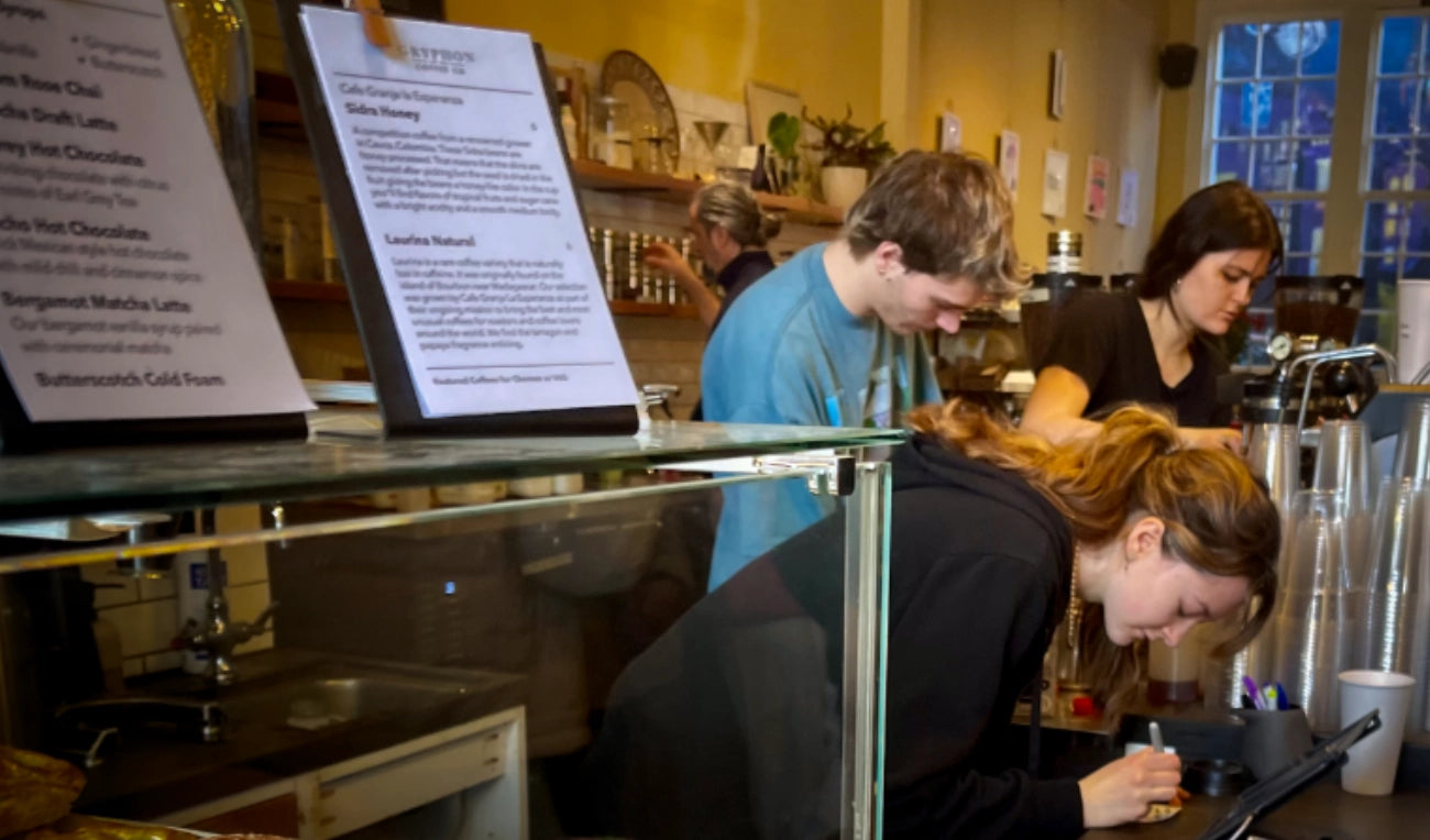 People working behind a counter in a coffee shop with a menu board in the foreground.