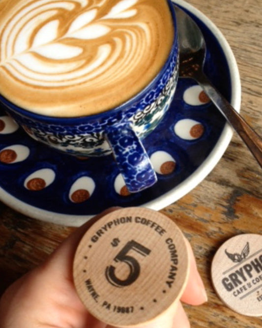 Cup of coffee with latte art on a saucer, accompanied by a wooden coin from Gryphon Coffee Company.